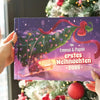 Weihnachtsbuch mit Titel „Emmas & Papas erstes Weihnachten 2025“ vor einem festlich dekorierten Tannenbaum.